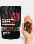Air-dried mackerel dog treats shown in a hand beside a 100g mackerel bites pouch for size comparison.