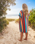 Person in a colorful striped hooded towel walking towards the beach from a sandy path lined with greenery.