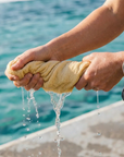 A close-up, of a swimmer’s hands wringing out a highly absorbent yellow chamois towel at an ocean pool. Water is shown streaming from the shammy cloth, demonstrating its quick-dry and reuse capabilities. In the background, a person in a blue swimsuit and goggles stands against sparkling turquoise water.