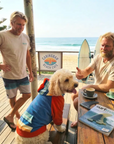 Two men with a dog wearing a dog hoodie at a beach cafe, with a surfboard and magazine on a table.