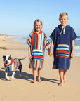 Two children and a dog wearing colorful beach hooded towels on a sandy beach in Australia with ocean in the background.