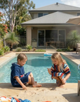 Two children wearing hooded towels, playing by a pool with a house and garden in the background