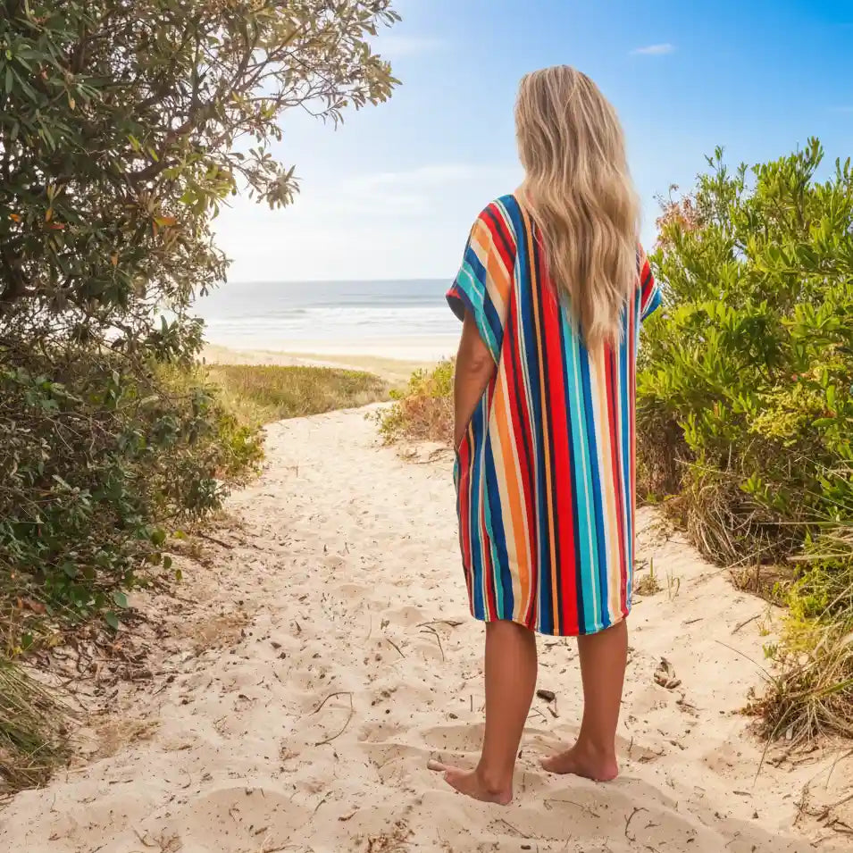 Person in a colorful striped hooded towel walking towards the beach from a sandy path lined with greenery.
