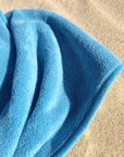 a blue dog towel laying on the sand at the beach, towel has a very soft looking texture.