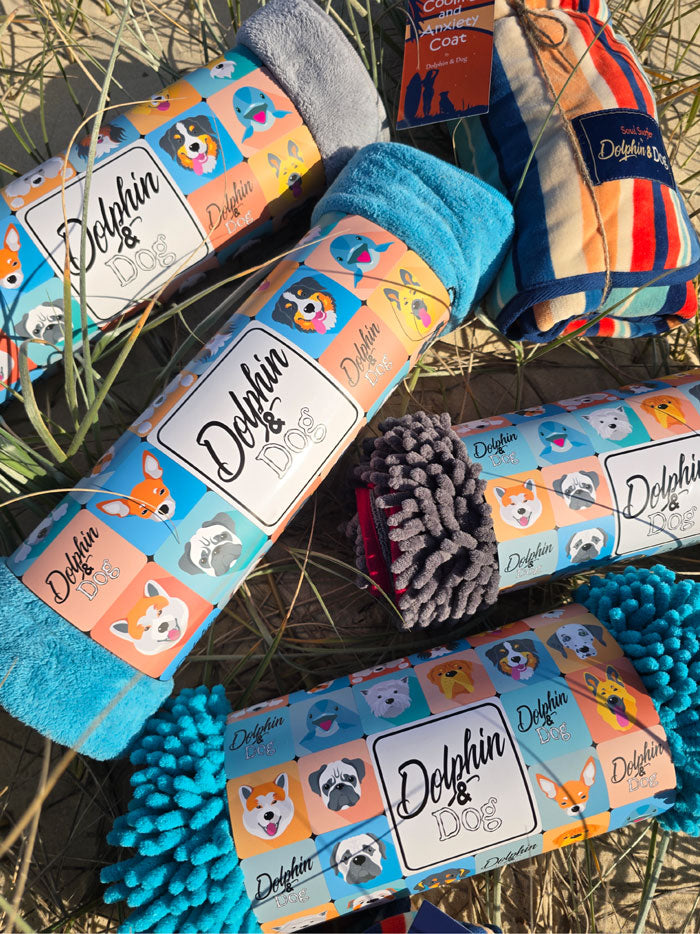 Dolphin and Dog, dog towels being displayed on the beach in colourful packaging, 3 different styles, dog bath towel, dog noodle towel and a dog wearable robe towel 