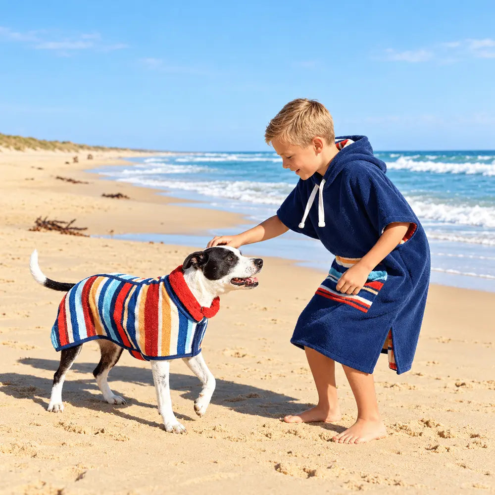 A boy and a dog on a sandy beach, with the boy wearing a navy blue hooded towel robe and the dog wearing a striped towel 
