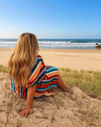 Woman sitting on a sandy beach wearing a colorful striped hooded towel, looking out at the ocean.