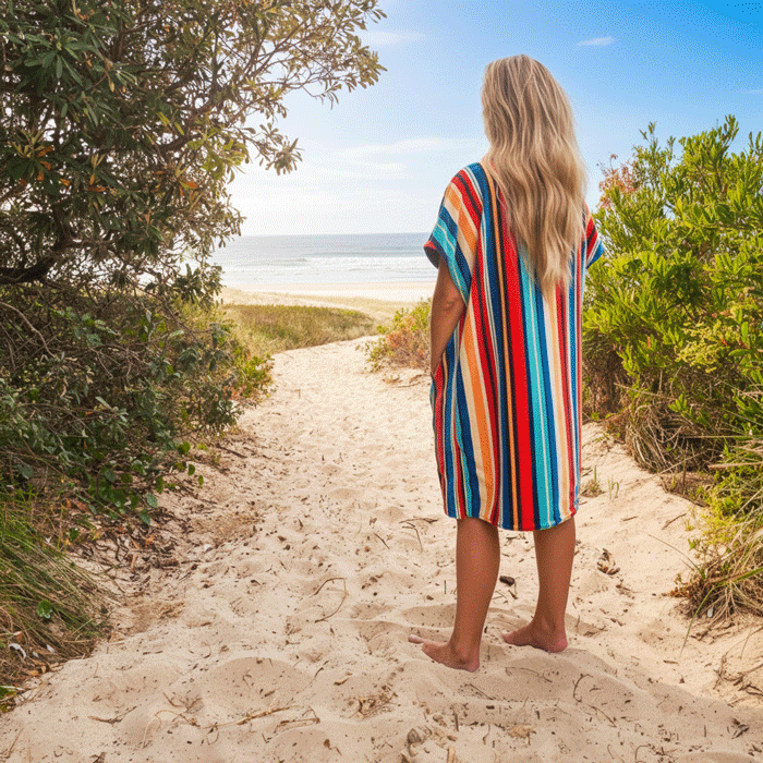 Person in a colorful striped hooded towel walking towards the beach from a sandy path lined with greenery.