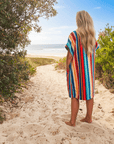 Person in a colorful striped hooded towel walking towards the beach from a sandy path lined with greenery.