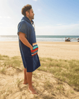 Man standing on a beach wearing a blue hooded towel with a colorful striped pattern.