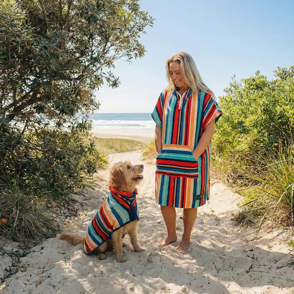 Woman and her dog at the beach wearing matching towels, dog is wearing a striped dog robe and the women a striped hooded towel 