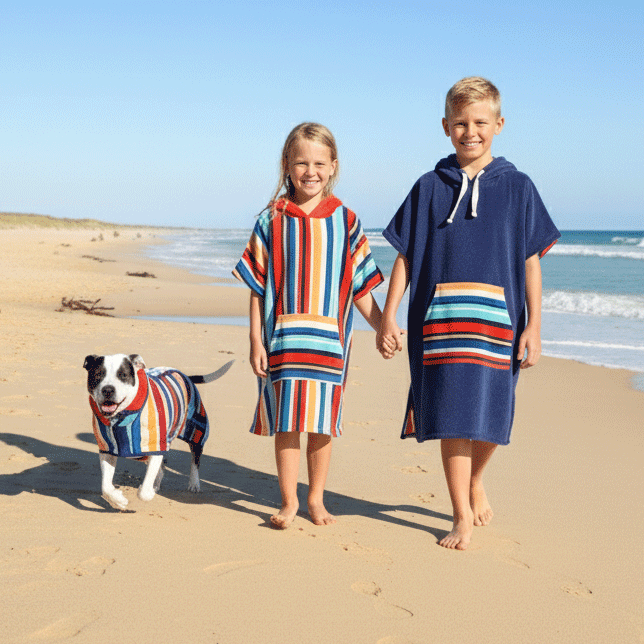 Two children and a dog wearing colorful beach hooded towels on a sandy beach with ocean in the background.