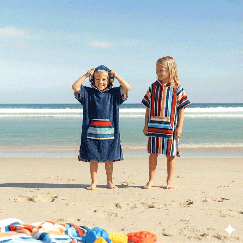 Two children on a beach wearing colorful kids hooded towels, with ocean and sky in the background
