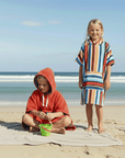 Two children on a beach wearing colorful hooded towels.