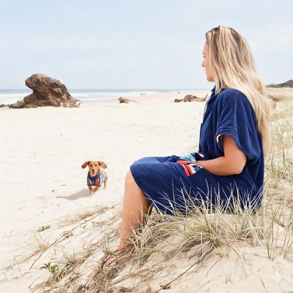 Woman in a blue hooded towel sitting on a sandy beach with a dog wearing a matching dog towel 
