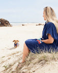 Woman in a blue hooded towel sitting on a sandy beach with a dog wearing a matching dog towel 