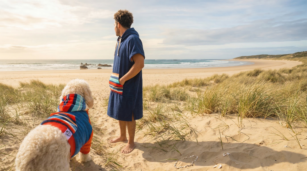 Man and dog on a sandy beach wearing matching clothes with grassy dunes and ocean in the background